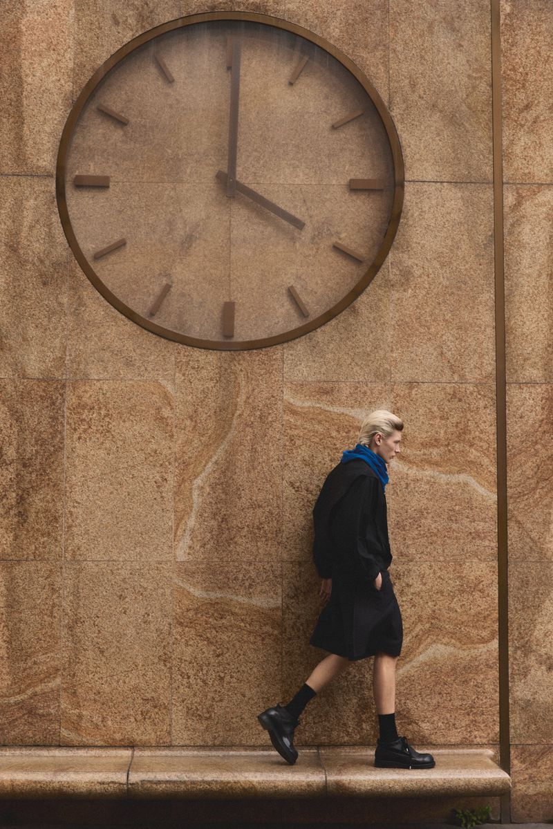 Model walking past a large wall clock on a textured stone building exterior for Numero Netherlands editorial