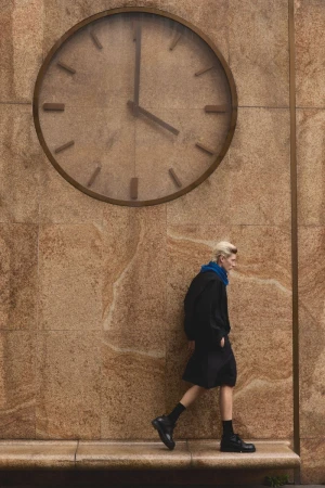 Model walking past a large wall clock on a textured stone building exterior for Numero Netherlands editorial