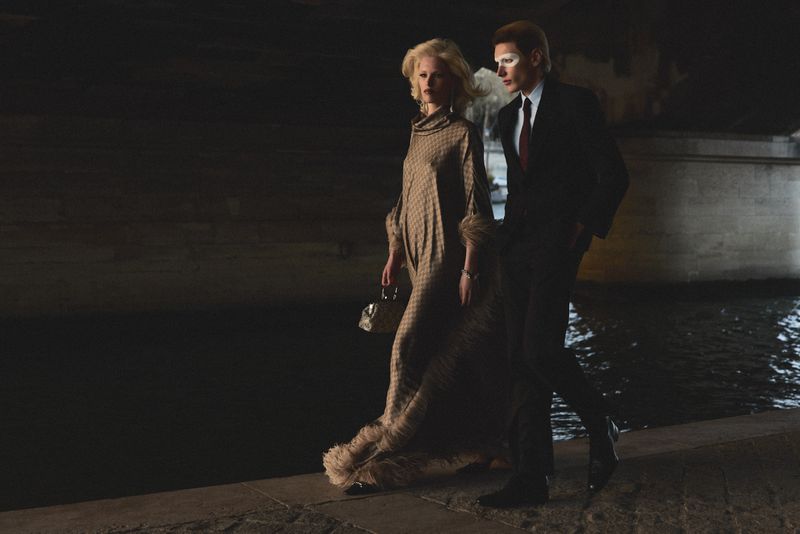 A blonde woman in a patterned dress and a man in a black suit walking along a stone embankment beside dark water.
