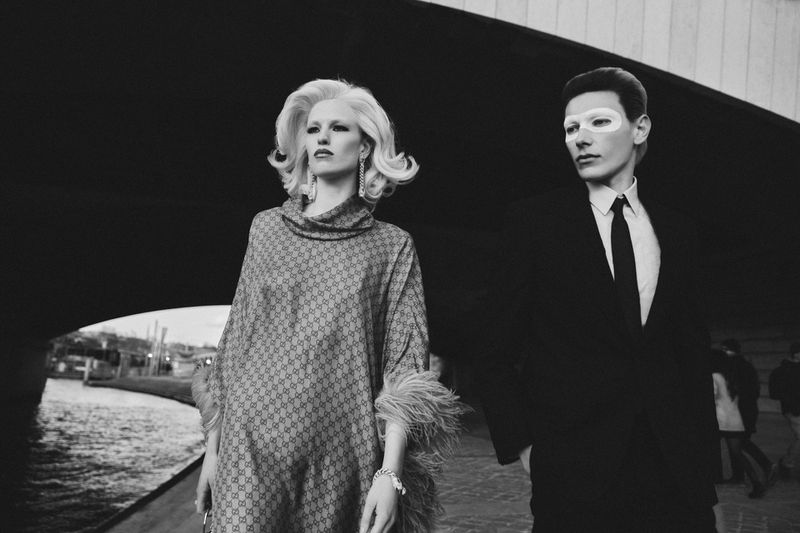 Black and white editorial portrait of two models standing outdoors near a bridge, wearing formal attire.