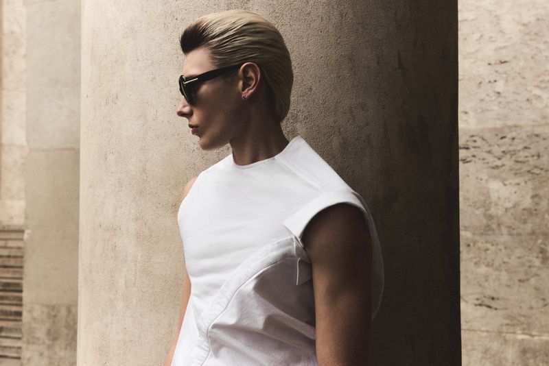 Profile portrait of a man wearing dark sunglasses and a white sleeveless top standing beside a stone column.