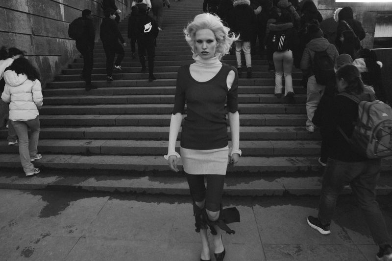 Model with blonde hair walking down public outdoor stone stairs surrounded by blurred crowds of pedestrians.