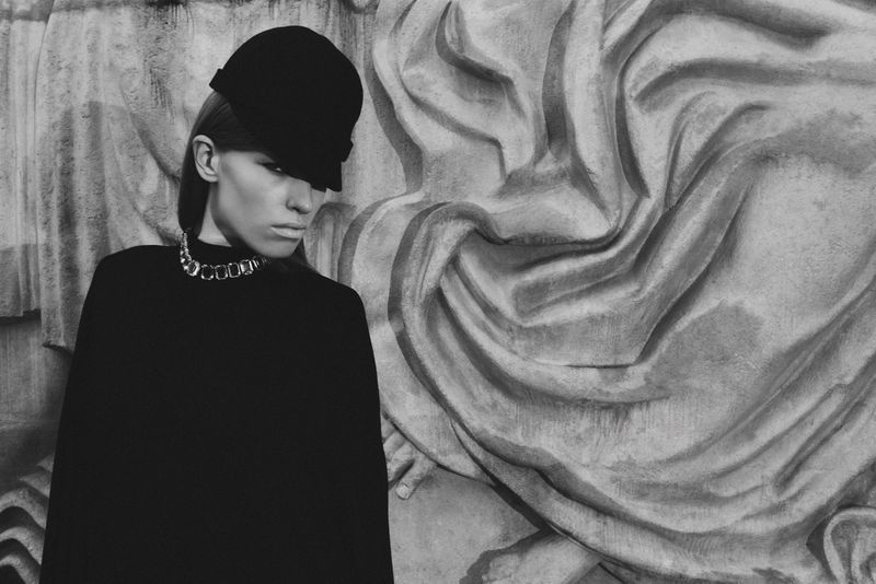 A solitary adult woman wearing a stylish hat and necklace poses against a stone architectural backdrop in a monochrome fashion photograph.