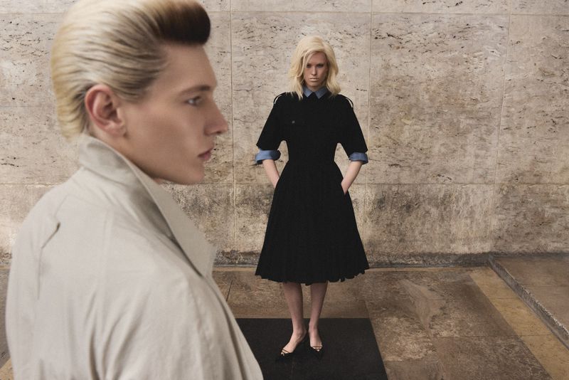 A blonde man in profile wearing a beige trench coat with a blonde woman in a black dress standing in the background.