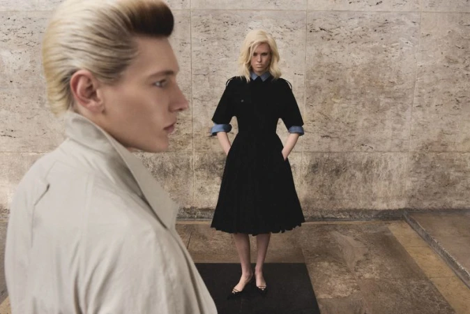 A blonde man in profile wearing a beige trench coat with a blonde woman in a black dress standing in the background.