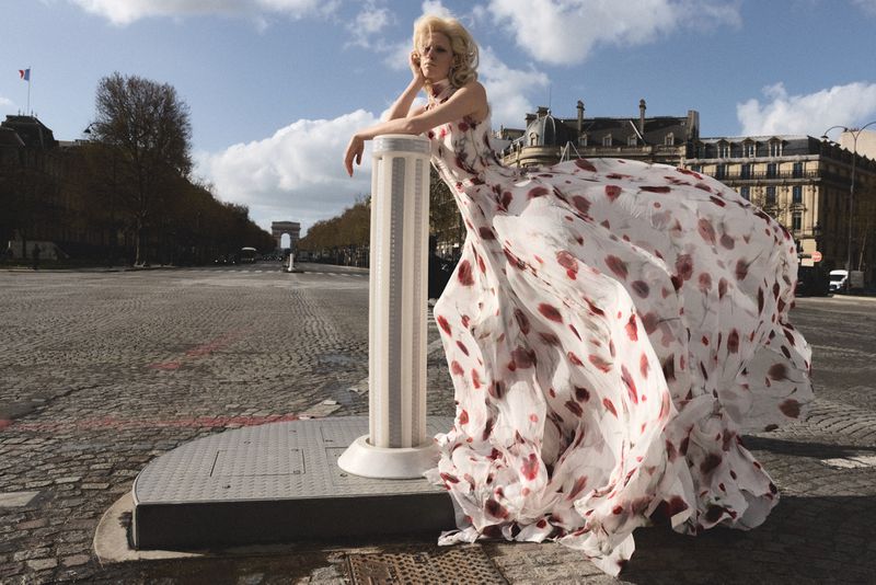 Model wearing a white floral print dress leaning against a pillar on a wide cobblestone street with a distant archway.