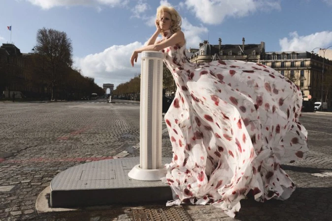 Model wearing a white floral print dress leaning against a pillar on a wide cobblestone street with a distant archway.