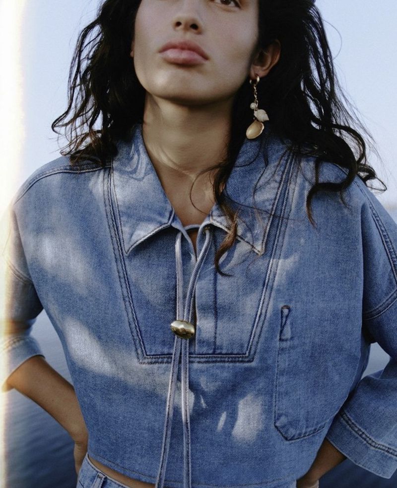 Model wearing a blue denim shirt with drawstring ties and shell earrings against an outdoor sky background