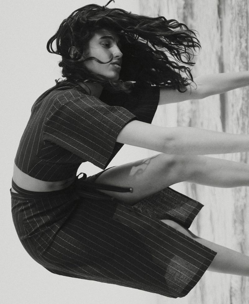 Black and white portrait of a woman with long dark wavy hair wearing a striped matching crop top and skirt in a studio setting.