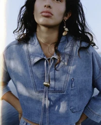 Model wearing a blue denim shirt with drawstring ties and shell earrings against an outdoor sky background