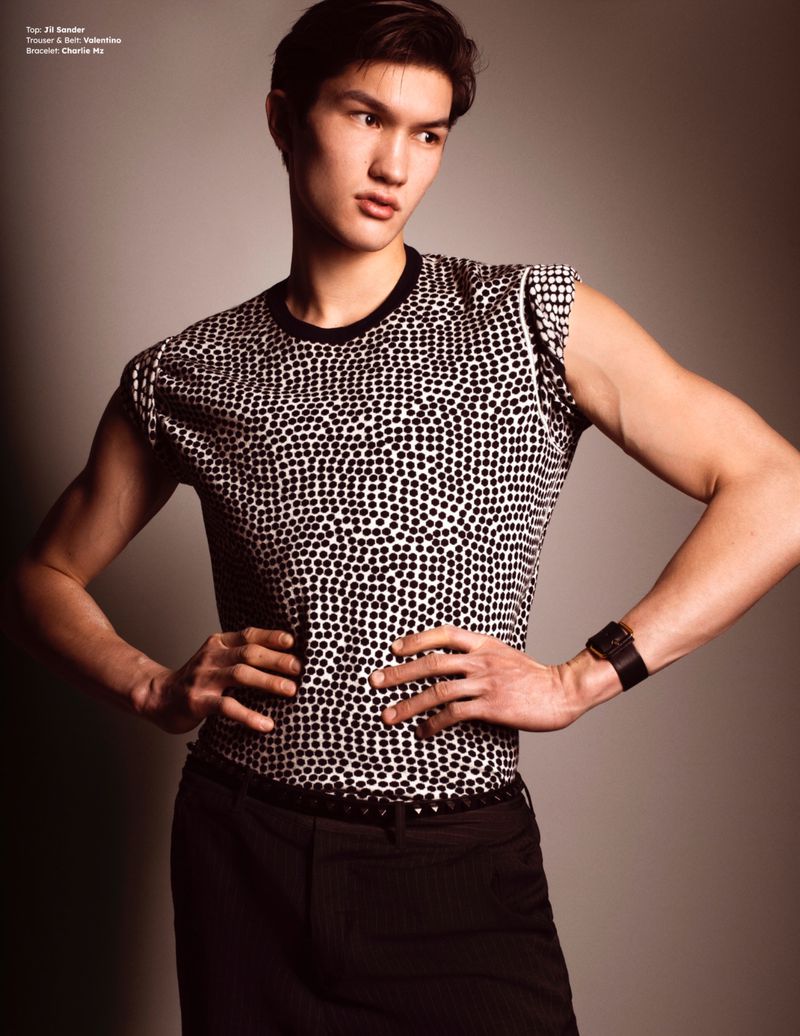 A male model posing with hands on hips wearing a patterned top and dark trousers against a neutral background.