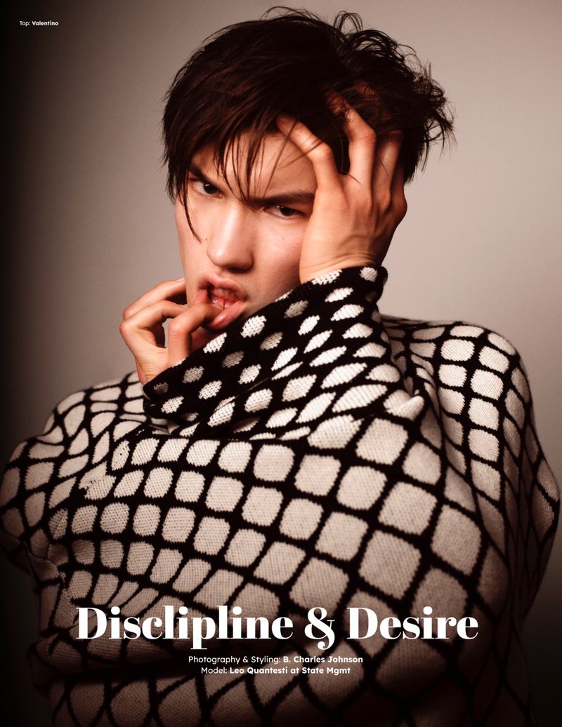 Male fashion model with hands in hair posing in a studio, wearing a black and white patterned sweater looking at the camera.