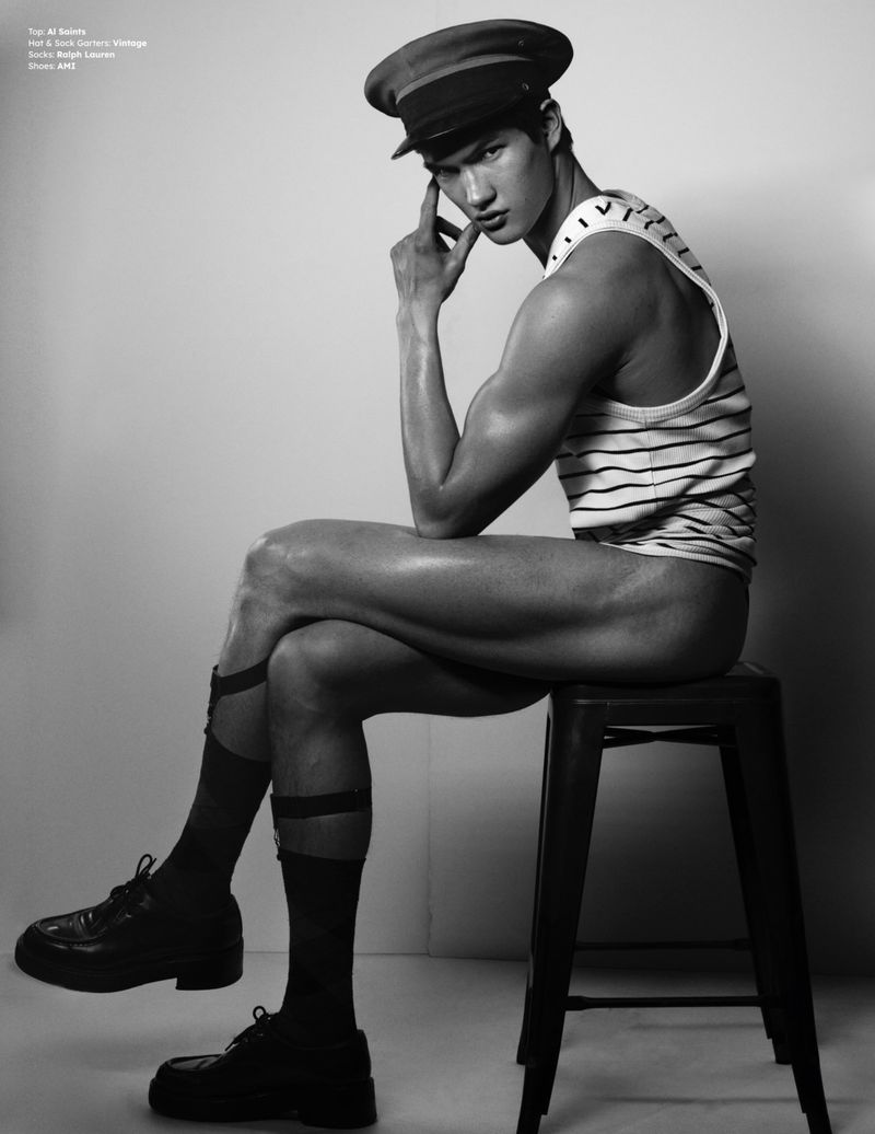 Monochrome fashion portrait of a male model wearing a striped tank top, cap, socks, and shoes, seated on a stool in a studio.