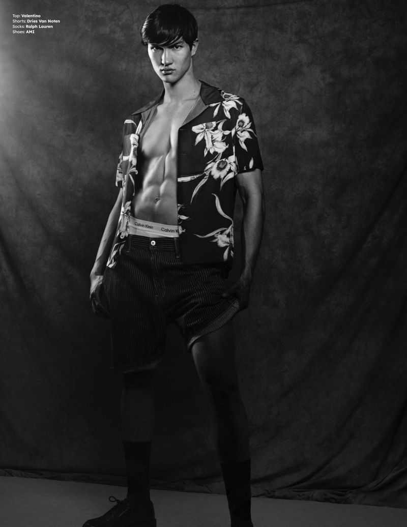 Portrait of a man wearing a patterned open shirt and dark shorts standing against a textured studio backdrop