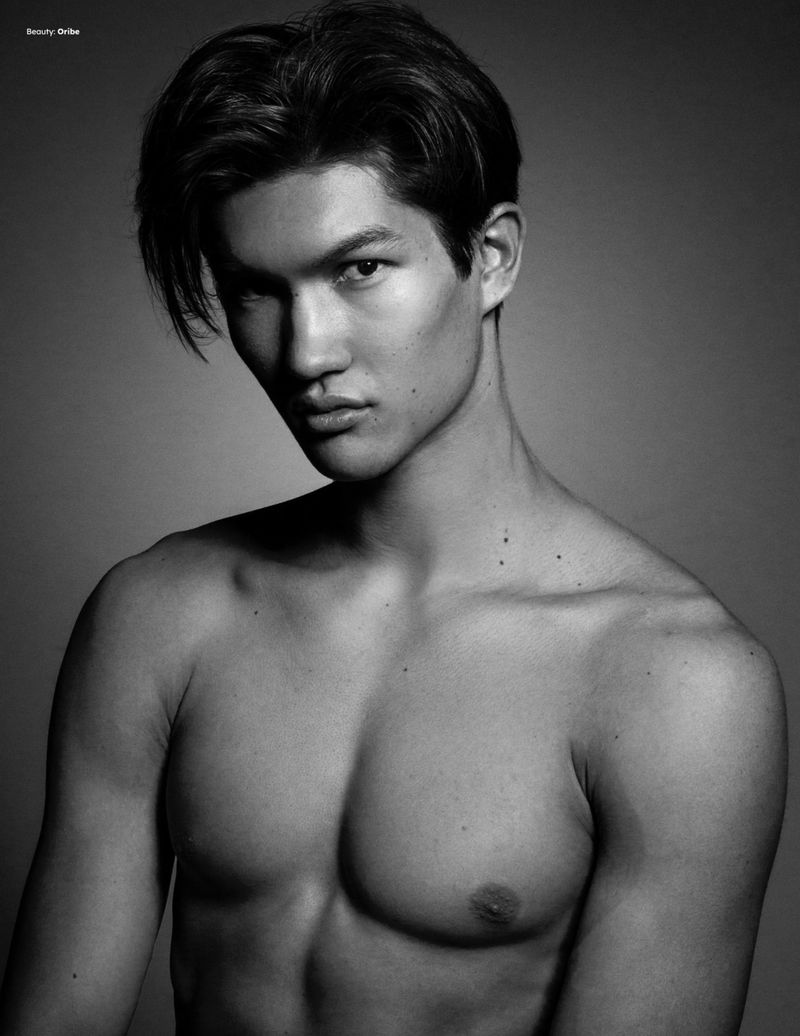 Monochrome portrait of a shirtless young man with dark styled hair looking intently at the camera against a grey gradient background