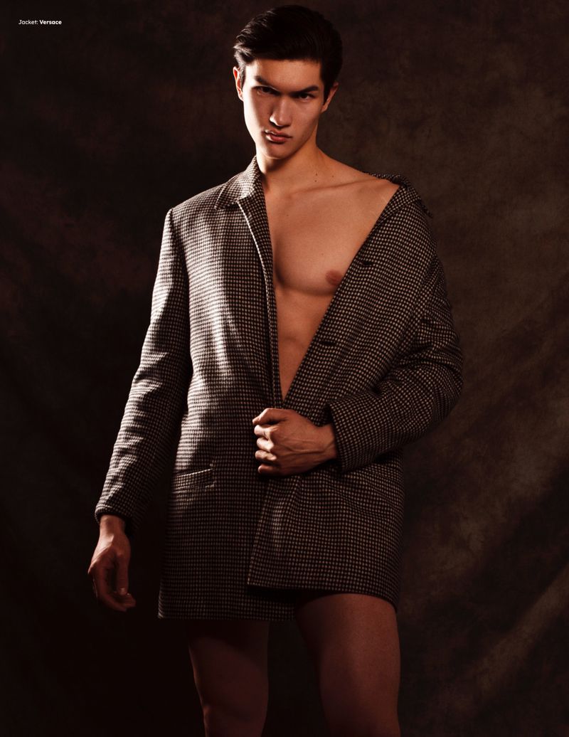 Man wearing a patterned oversized blazer unbuttoned over bare chest against dark textured background