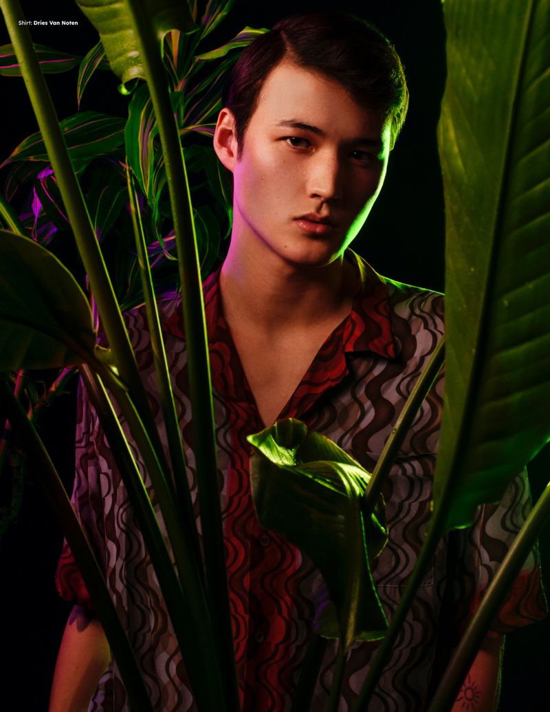 Close-up portrait of a man in a patterned short-sleeve shirt partially framed by large green tropical leaves against a dark background.