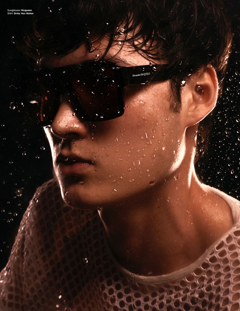 Close-up portrait of a young man wearing dark sunglasses and a mesh shirt, covered in water droplets against a dark background.