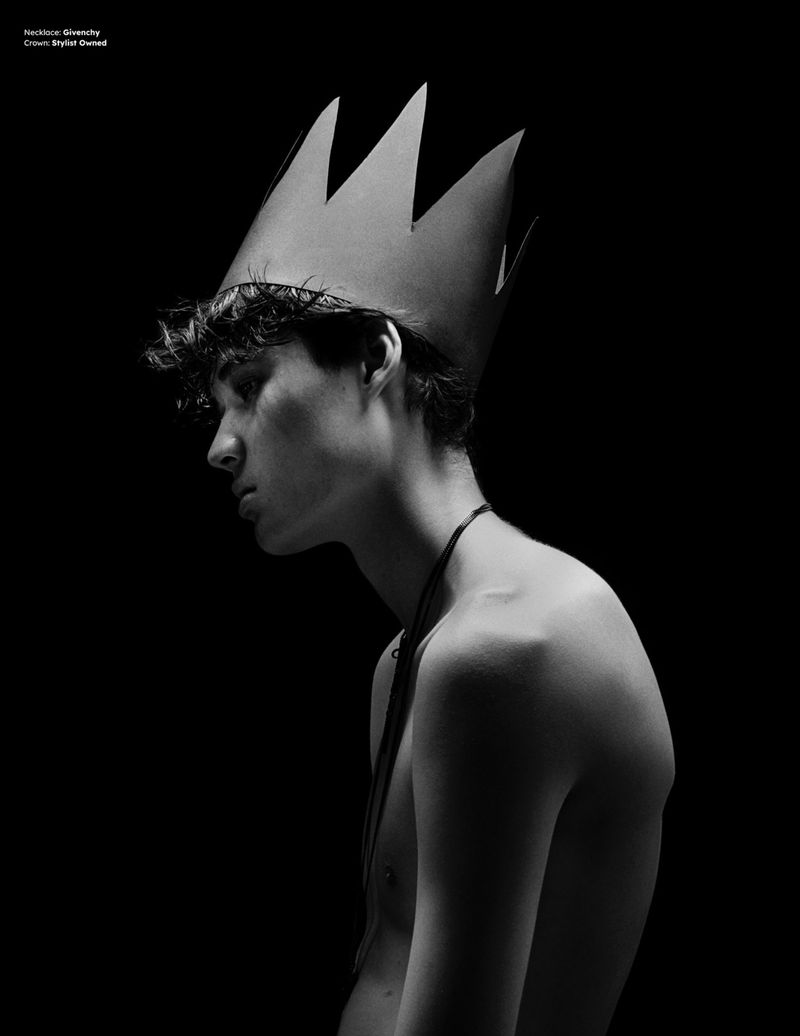 Monochrome portrait of a young man wearing a pointed paper crown and a thin necklace against a black background.