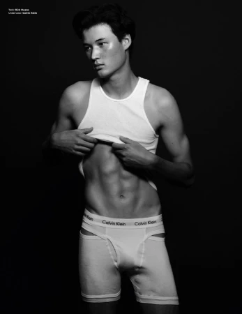 Monochrome portrait of a young man wearing white underwear and a white tank top against a dark background.
