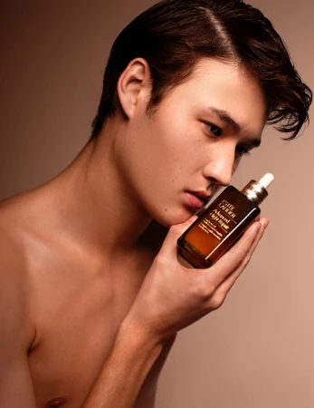 Close-up portrait of a young man holding a brown serum bottle against a warm neutral background for Desnudo Magazine.