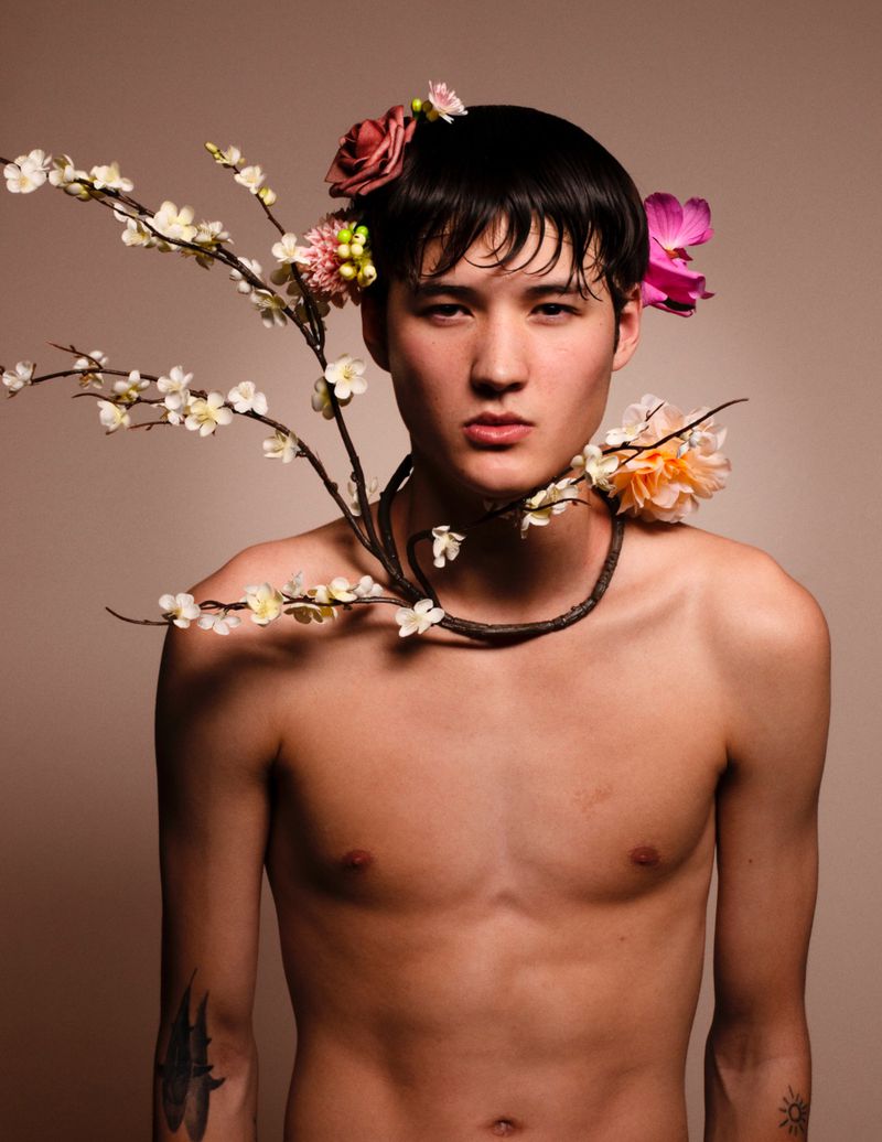 Shirtless man with dark wet hair wearing floral branch necklace and flowers in hair against neutral background
