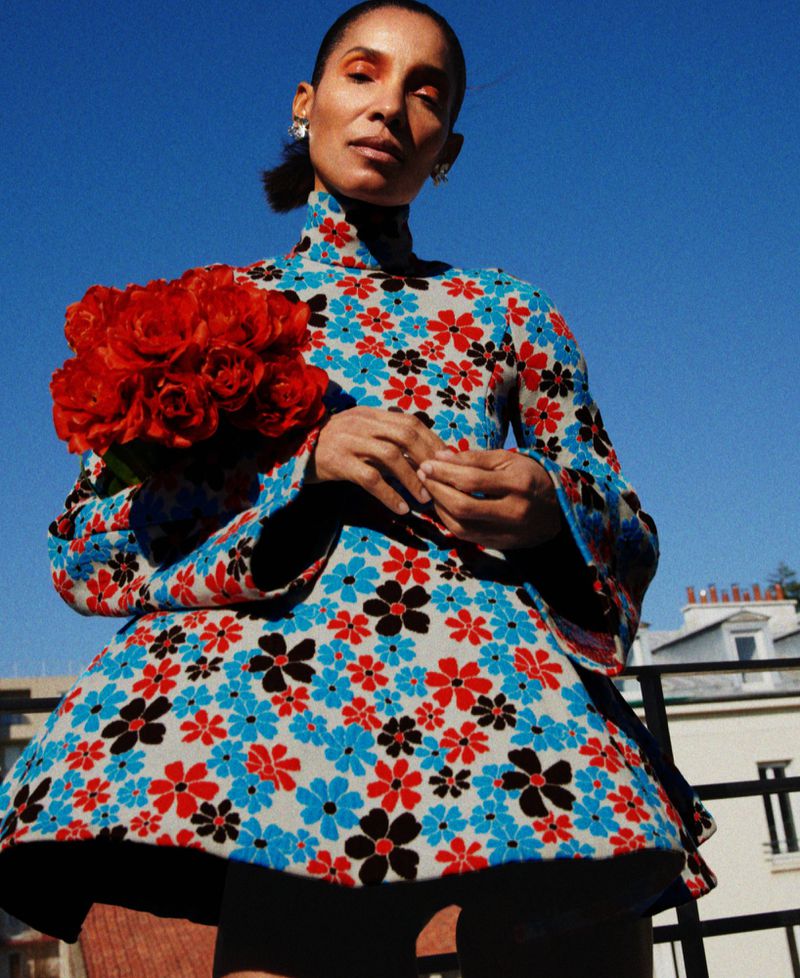 Model in a floral patterned dress holding a bouquet of red roses against a clear blue sky background