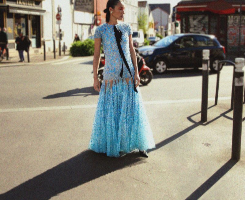 Fashion shoot in urban environment: A woman in a light blue gown with a black sash stands on a city street during daylight hours.