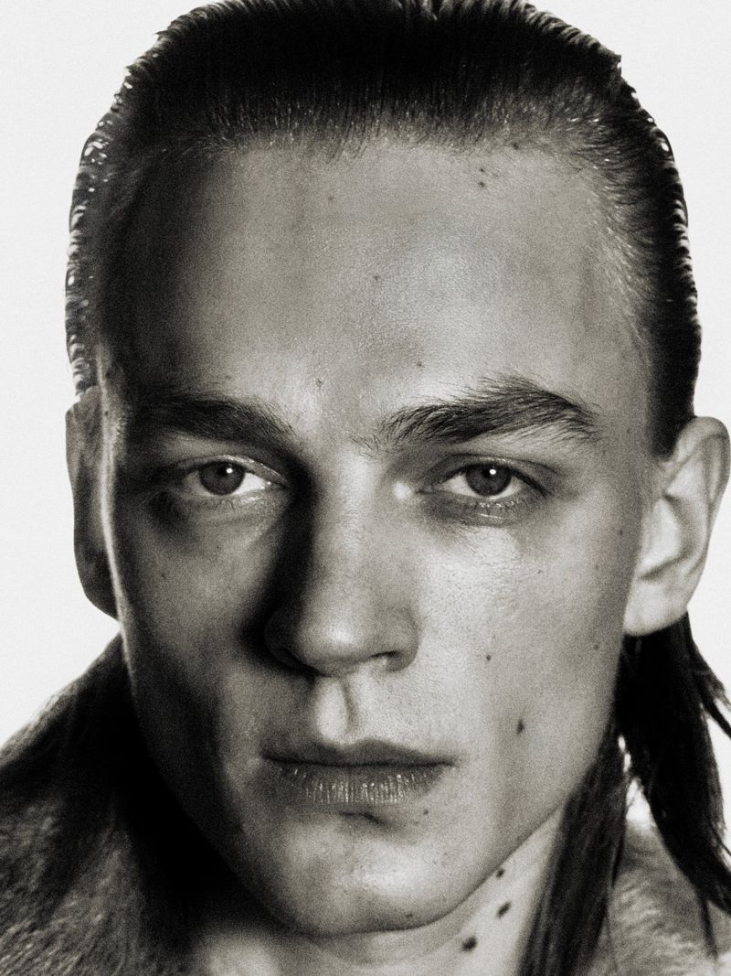 Close-up monochrome portrait of a young man with slicked-back hair and a textured collar against a bright white background.