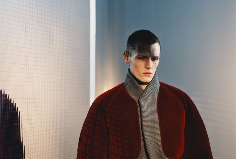 Close-up portrait of a male model wearing a red structured jacket over a grey ribbed turtleneck sweater.