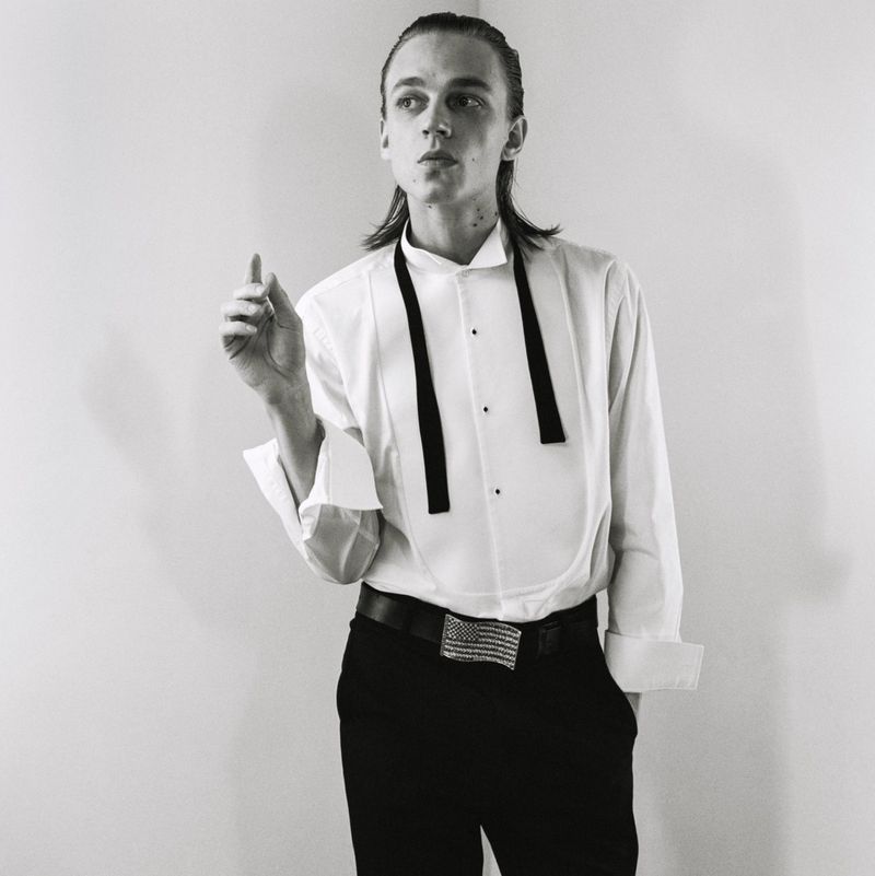 Monochrome portrait of a male model in a white tuxedo shirt and dark trousers against a plain studio background.