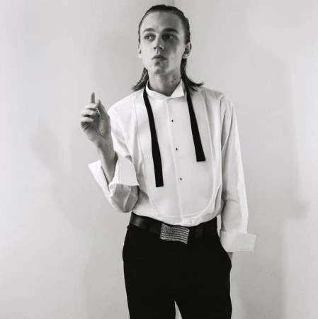 Monochrome portrait of a male model in a white tuxedo shirt and dark trousers against a plain studio background.