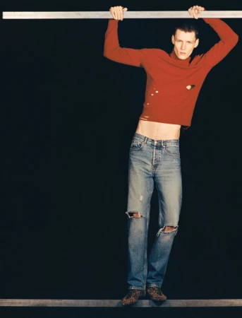 Model in a red long-sleeve top and distressed blue jeans hanging from a horizontal bar against a dark background