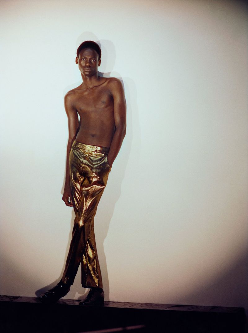 Shirtless black model wearing metallic gold trousers standing against a white wall with soft shadow