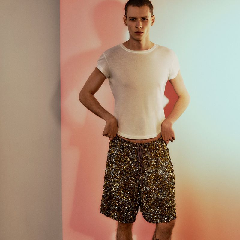 Model wearing a white t-shirt and sequin shorts standing against a gradient pink and blue background for Public Editions.