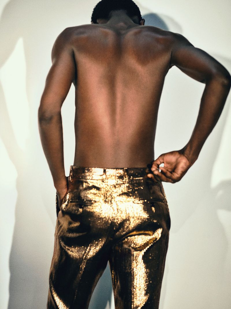 Shirtless male model from behind wearing shiny metallic gold trousers against a white seamless background