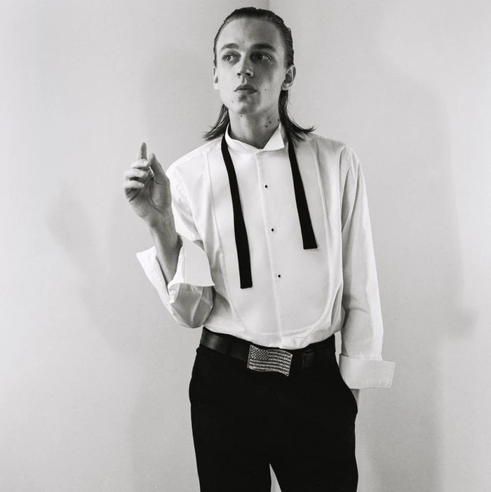 Monochrome portrait of a male model in a white tuxedo shirt and dark trousers against a plain studio background.