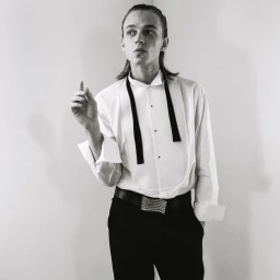Monochrome portrait of a male model in a white tuxedo shirt and dark trousers against a plain studio background.