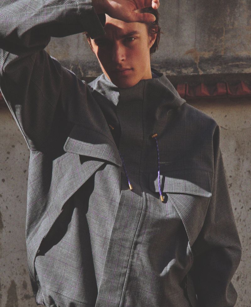 Young man wearing a patterned grey hooded jacket holding his hand to his forehead in harsh sunlight.
