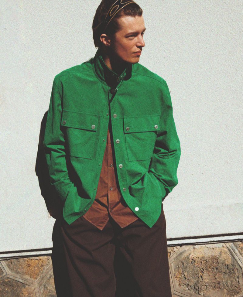 Man wearing a green jacket over a brown shirt leaning against a light textured wall outdoors.