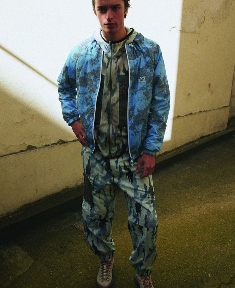 Young man standing outdoors wearing a blue camouflage jacket and matching trousers for M Le magazine du Monde