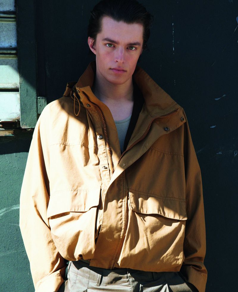 Man in a caramel-colored jacket stands against a dark backdrop, showcasing casual fashion and natural lighting in an outdoor setting.