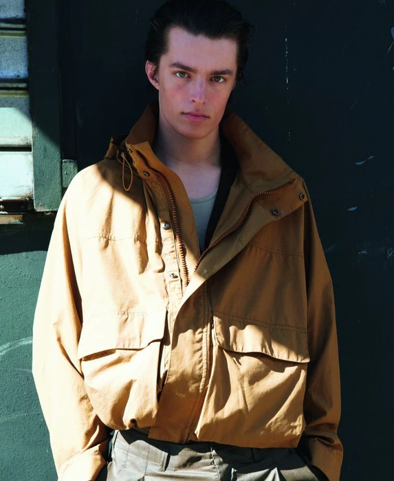 Young man wearing a camel hooded jacket standing outdoors against a dark wall for M Le magazine du Monde