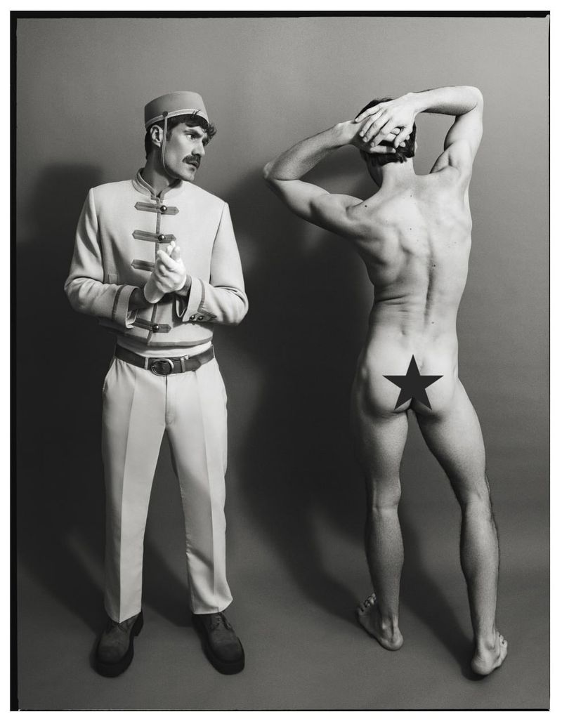 A hotel bellhop in a uniform looking at a shirtless man standing with his back turned, featuring a star graphic on his hip.