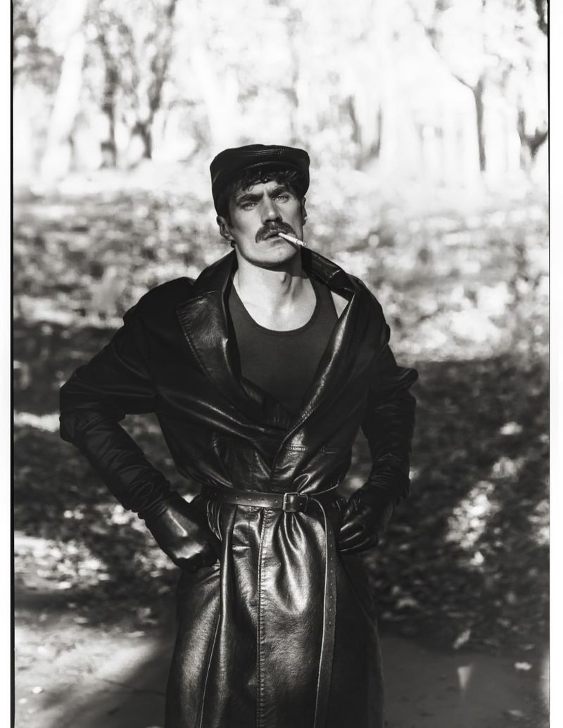 Monochrome portrait of a man with a mustache wearing a leather coat, cap, and gloves outdoors in a wooded setting.