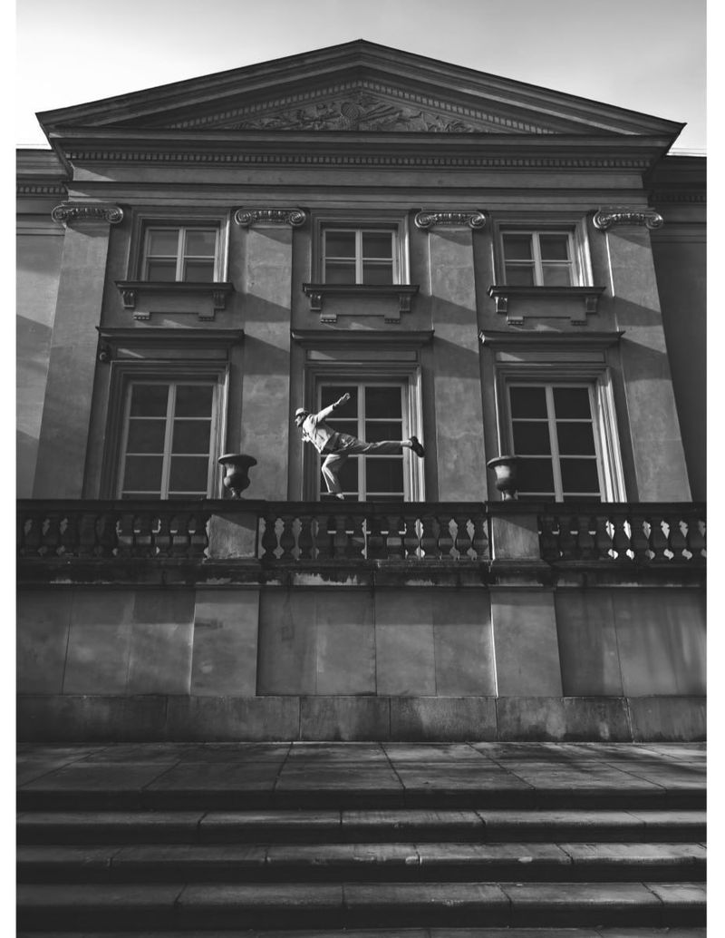 Monochrome architectural photograph of a person balancing on a stone balcony railing of a classical building.