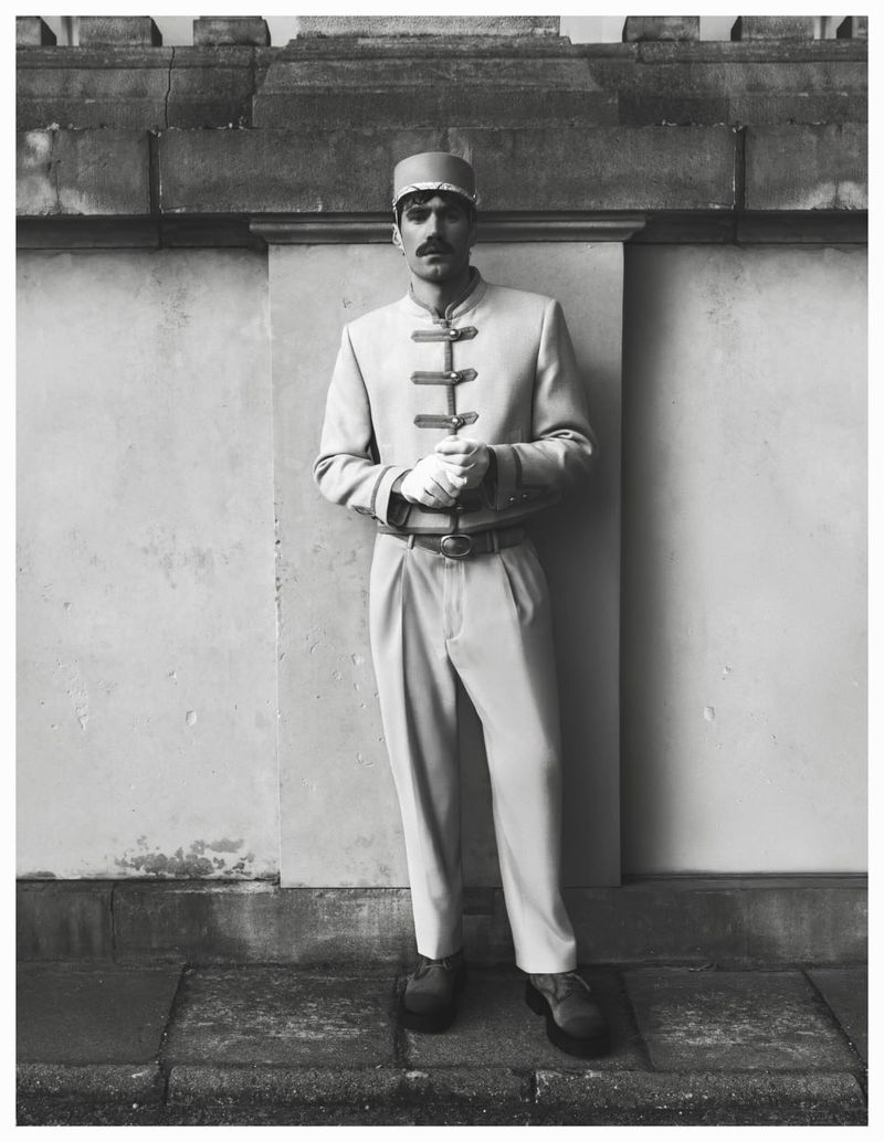 Man wearing a service uniform with high-button jacket and cap posing against a stone wall for Behind the Blinds.