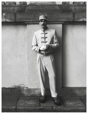 Man wearing a service uniform with high-button jacket and cap posing against a stone wall for Behind the Blinds.