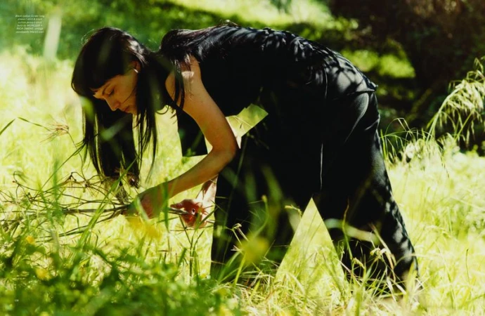 Woman with long dark hair wearing a black outfit bending over in a grassy field for Pop Magazine editorial shoot.