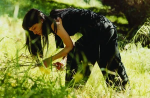Woman with long dark hair wearing a black outfit bending over in a grassy field for Pop Magazine editorial shoot.